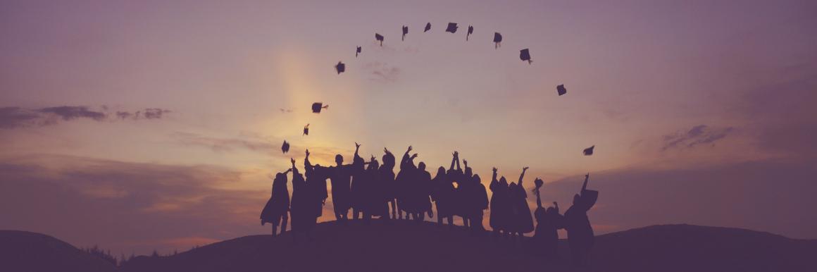 A group of graduates at dusk throwing their caps into the air, forming an arch of hats over the line of people