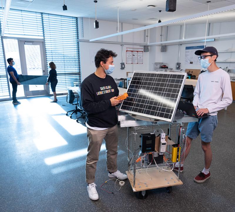 Two students wearing face masks, separated by a solar panel, in a discussion.