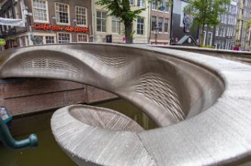 Side view of a highly curved, 3D-printed, steel, pedestrian bridge spanning over a water canal in Amsterdam.  Thin layers are visible in the bridge showing where beads of steel weld material were laid down to form the bridge.
