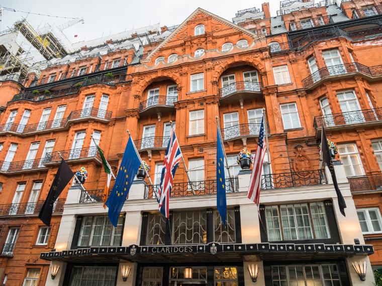 The front of a six-story, historic brick hotel with significant iron scrollwork and brick detail. Numerous flags, including Great Britain, USA, and the European Union, fly from the building.