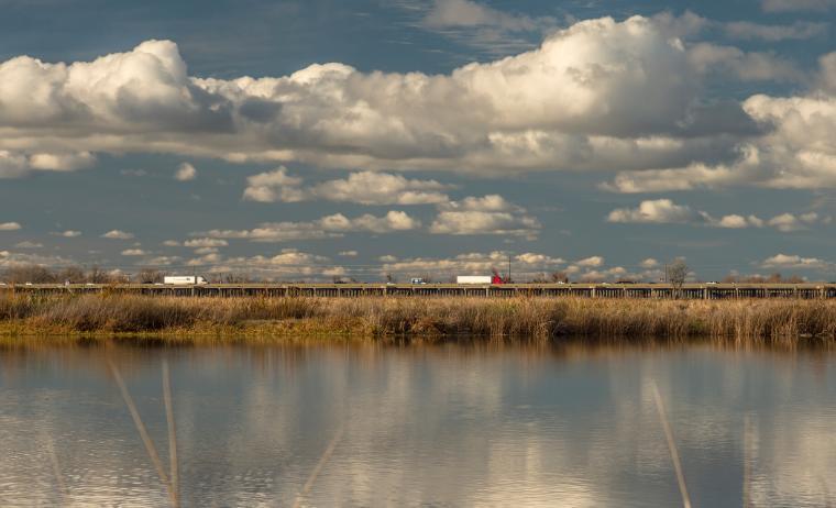 Large cumulus clouds are reflected in the waters of a flooded field.  The far edge of the floodplain is filled with reed grass and an elevated highway is shown in the background keeping cars and semis above the floodplain.