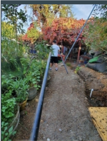 gravel pathway lined with green shrubbery on the left side of the photo. A male student, standing in the background, wearing black shorts and a blue shirt is hold a long, skinny pipe in his right hand, and a longer pipe in his left hand. 