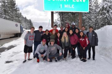 A group photo of 18 students in the snow in front of the Donner Lake 1 mile highway sign