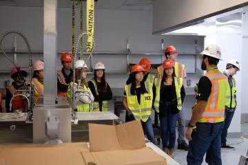 A group of 9 students wearing hard hats and yellow vests in a warehouse for a general contractor.