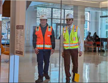 Two male CESE students from SCU wearing hard hats and safety vests walk in the Sobrato Campus of Discovery and Innovation.  The man wearing the orange safety vest is of Asian descent and the man wearing the yellow safety vest is of Hispanic descent. 
