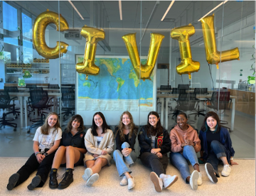 Seven female CESE students sit outside the geotechnical engineering laboratory below balloons spelling civil. A map showing global tectonic plates is visible.  Students are of White, Black, Asian, and Hispanic descent. 
