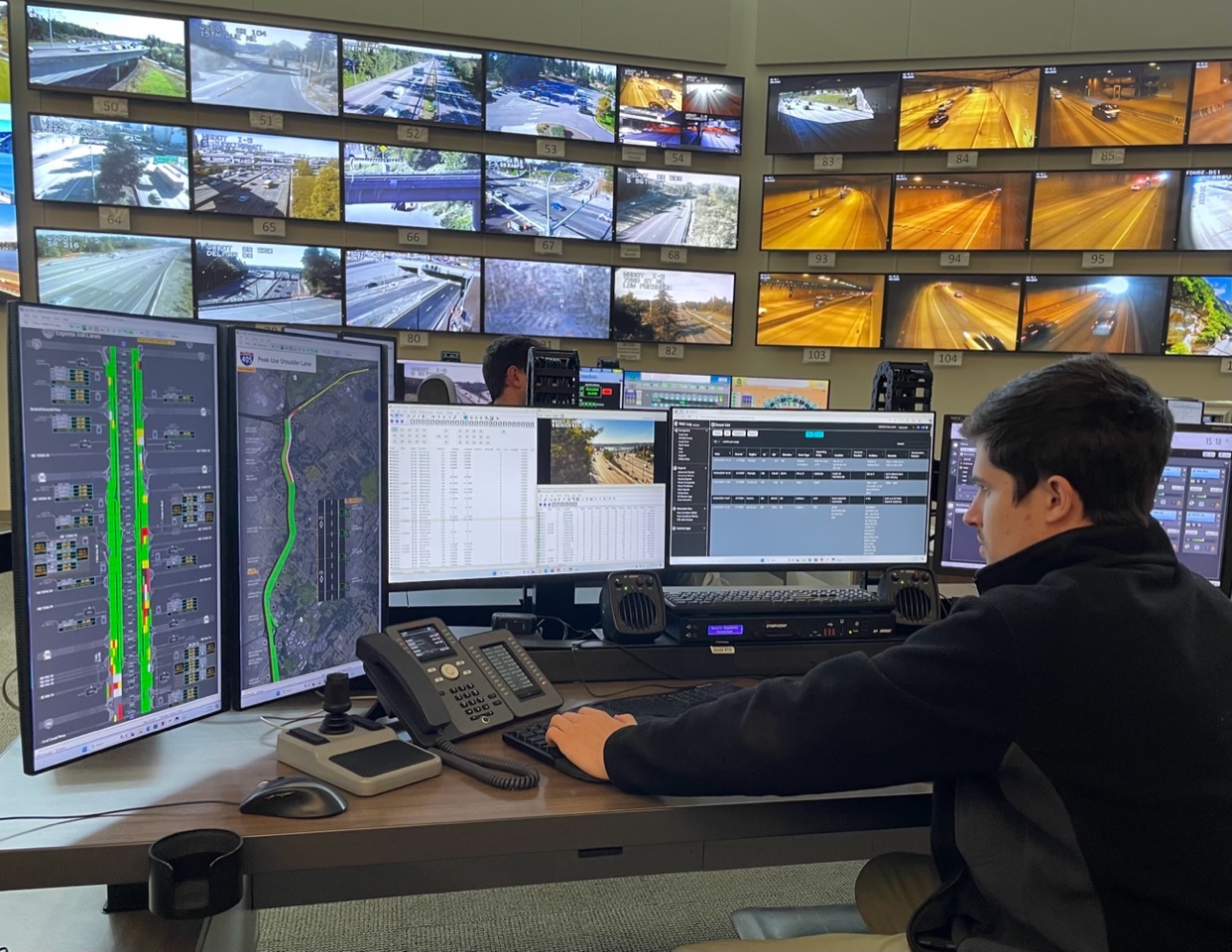 Male sitting at a desk in an office working on five monitors and in the background there is a wall of tv monitors displaying camera footage.