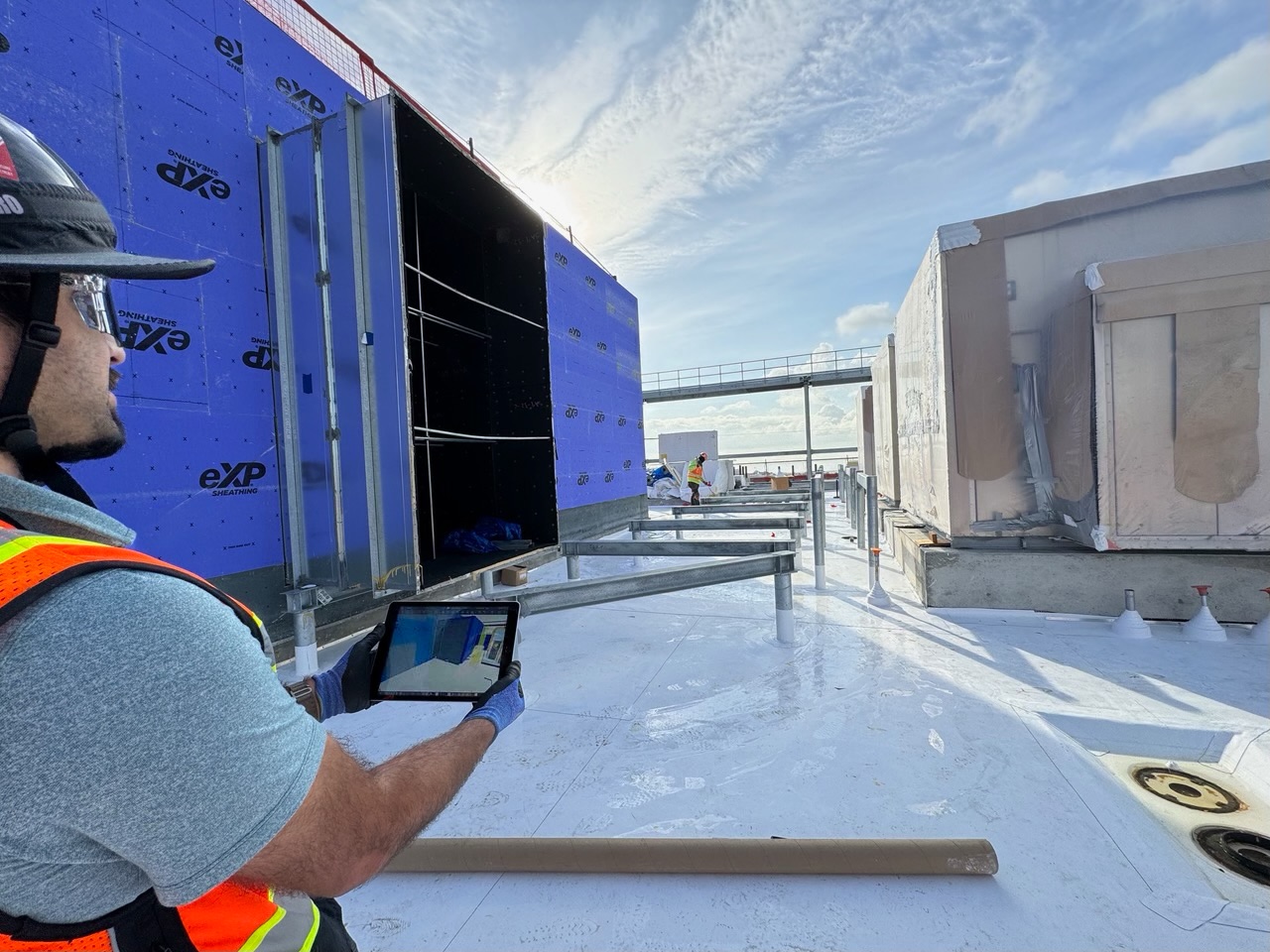Male wearing an orange construction vest reviews a 3D model on an iPad on an under construction rooftop.