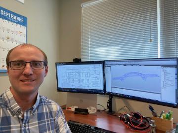 Photo of a male sitting at a desk with two computer monitors behind him displaying structural calculations and drawings for a new bridge design.