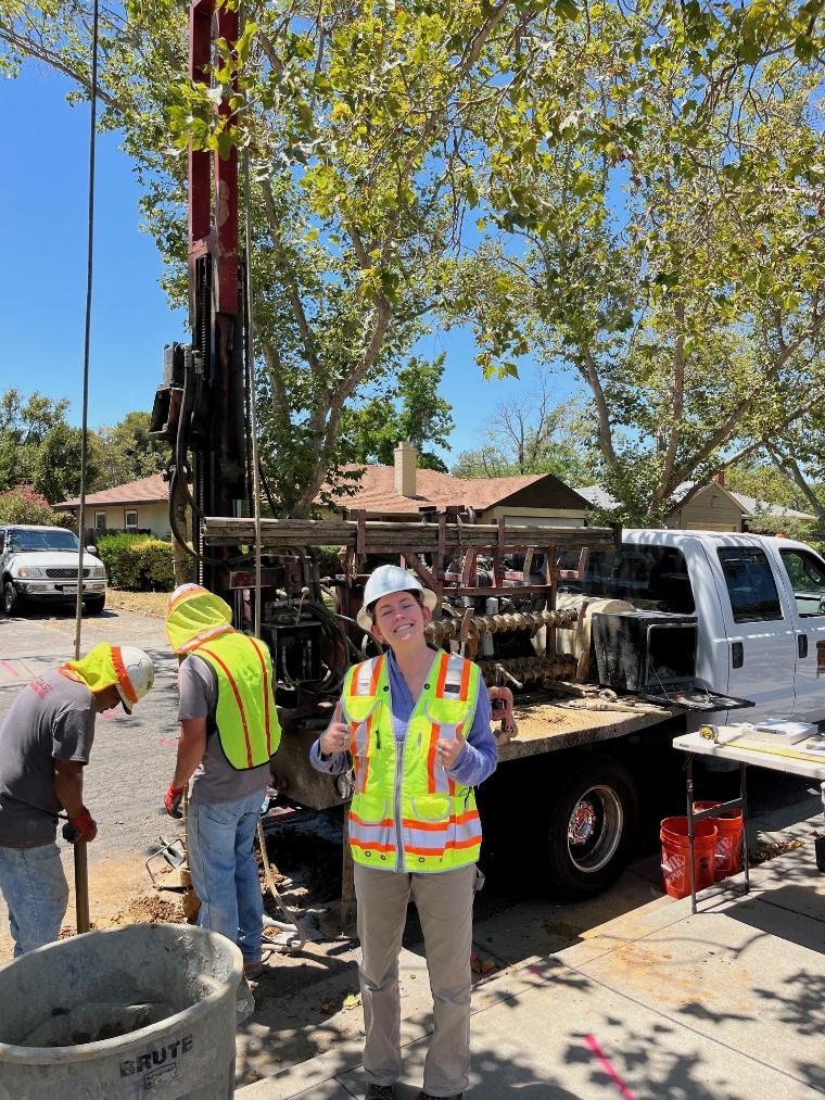 Civil engineering student wearing a white hard hat and construction vest while observing geotechnical drilling for a sanitary sewer replacement.
