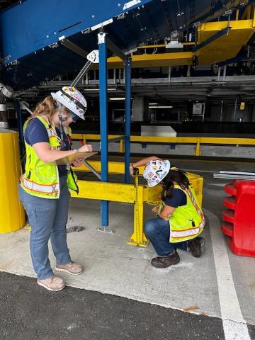 Two women civil engineers wearing hard hats and construction vests, one looking at plans and the other measuring a structure.