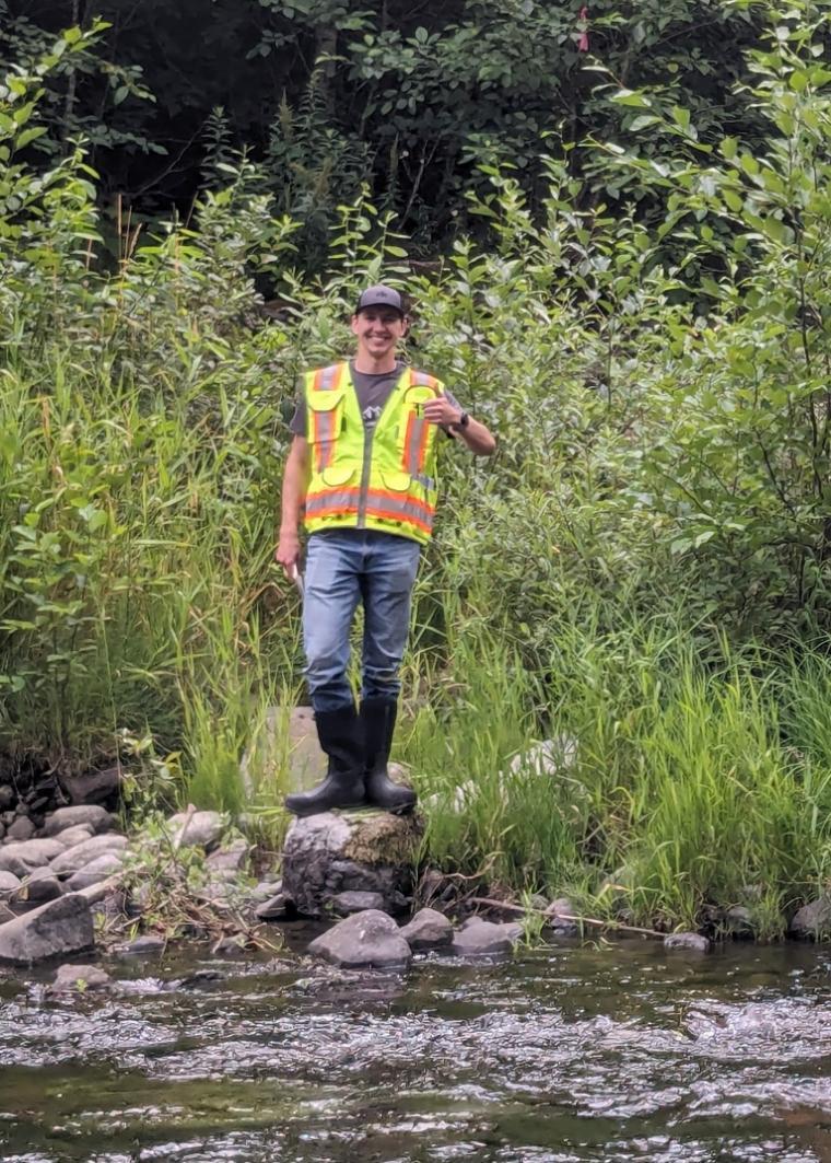 Man in a yellow construction vest standing on a rock beside a river.