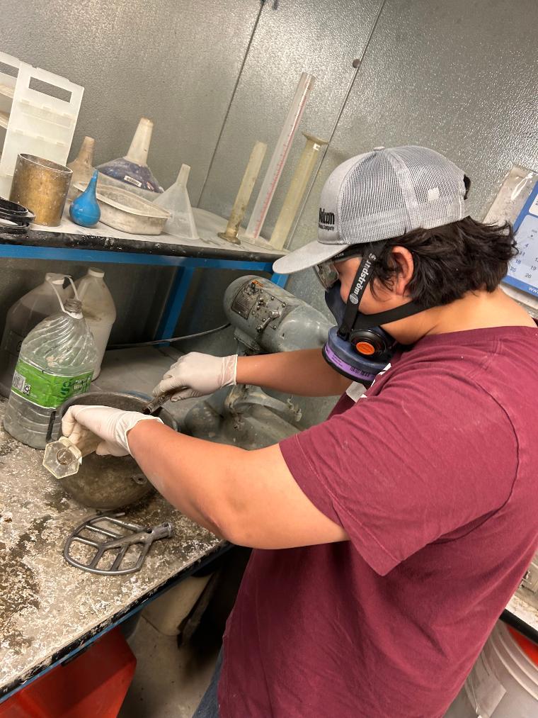 Student wearing a red shirt, grey baseball cap, and safety gas mask testing mixes for greener concrete on a work bench.