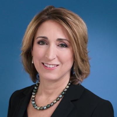 Person smiling in front of a blue background wearing a black blazer.