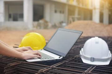 Hands typing on a laptop computer, with hard hats and construction site in the background image link to story