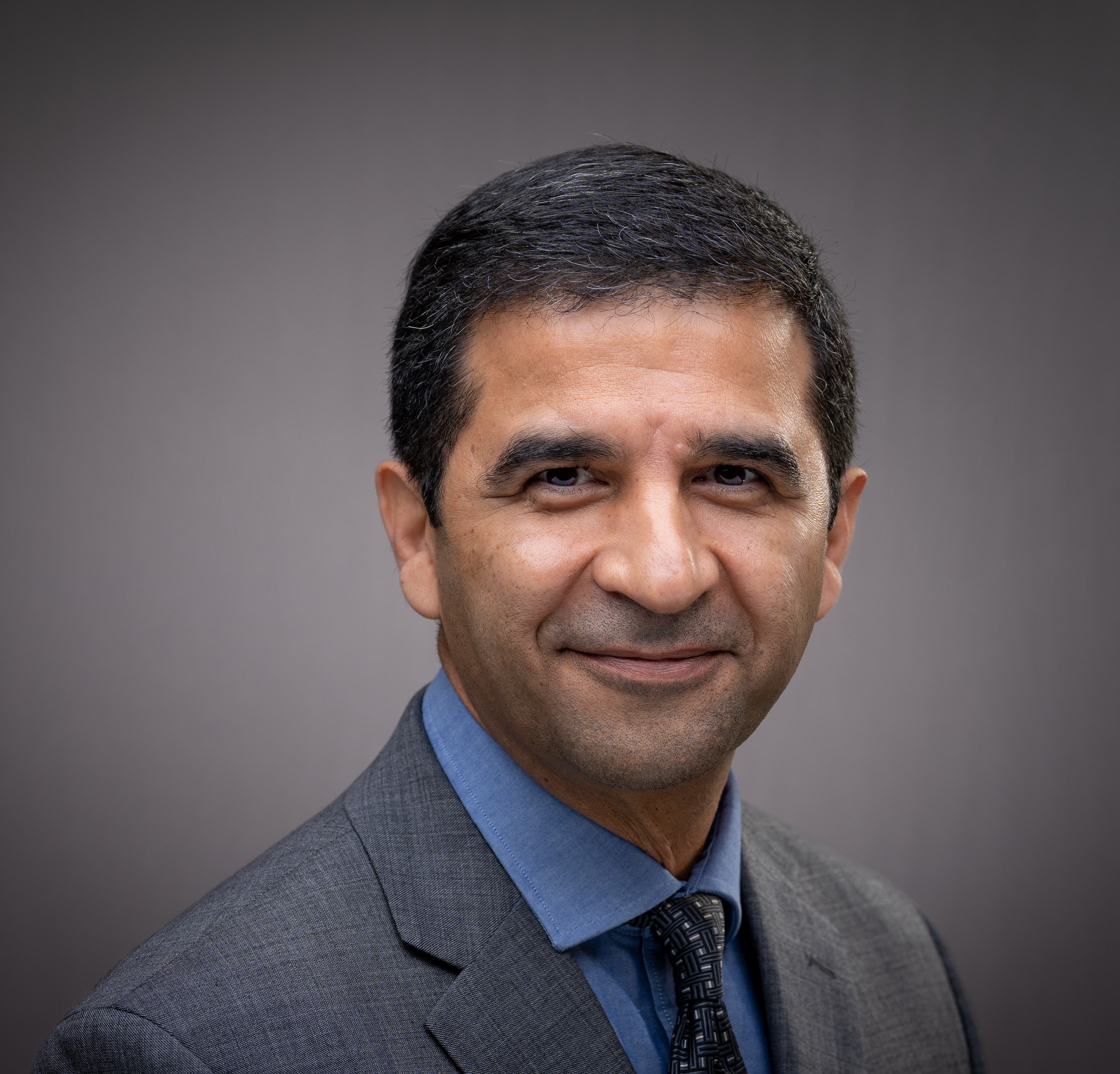 A man in a suit smiling against a grey background.