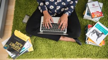 Student on a laptop sitting on a lawn with books all around 