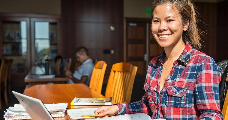 A female student studying with her laptop and books, the sun shining on her face.