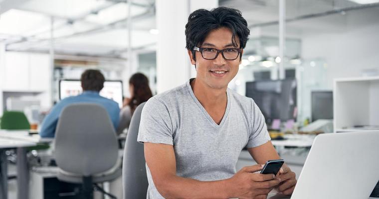 Man sitting at his laptop smiling at the camera in an office setting. image link to story