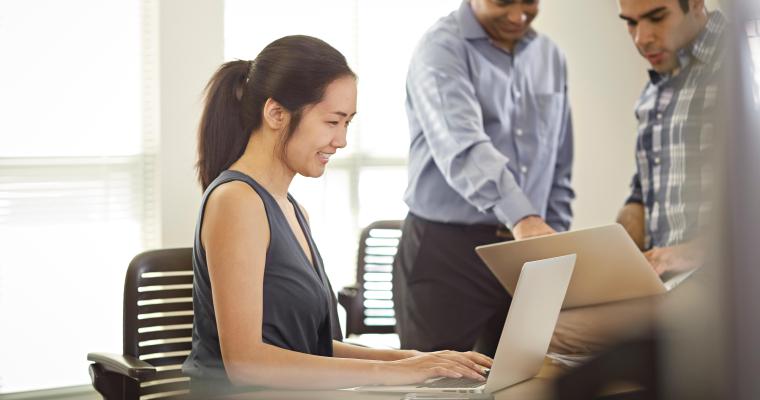 Female student sitting at a laptop with a professor and fellow student sanding nearby.